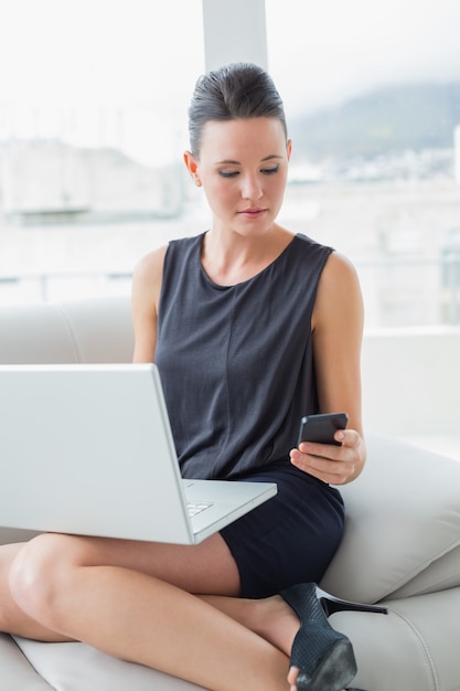 Belle femme bien habillée en utilisant un ordinateur portable et un téléphone portable sur le canapé