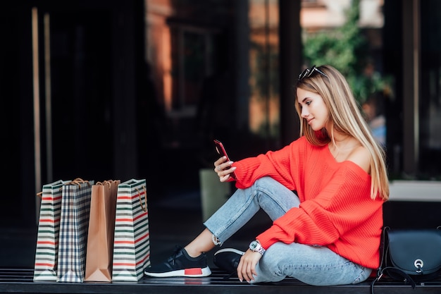 Belle femme blonde assise dans la rue et entourée de sacs à provisions et regardant au téléphone.