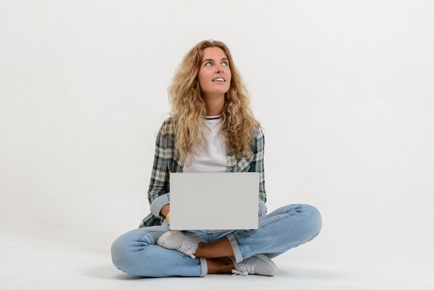 Belle femme blonde assise sur le sol avec un ordinateur portable sur le mur blanc