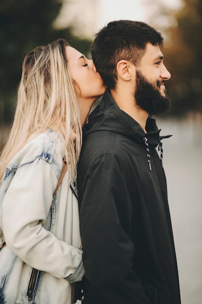 Belle femme blonde aux cheveux longs embrassant la tête avec les yeux fermés de son petit ami avec amour pendant qu'il regarde ailleurs en souriant heureux en marchant le matin.