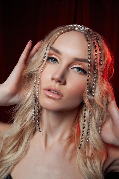 Belle femme blonde aux cheveux longs sur fond de velours rouge Bijoux de cheveux sur une tête de femme maquillage moderne et flèches sur les yeux