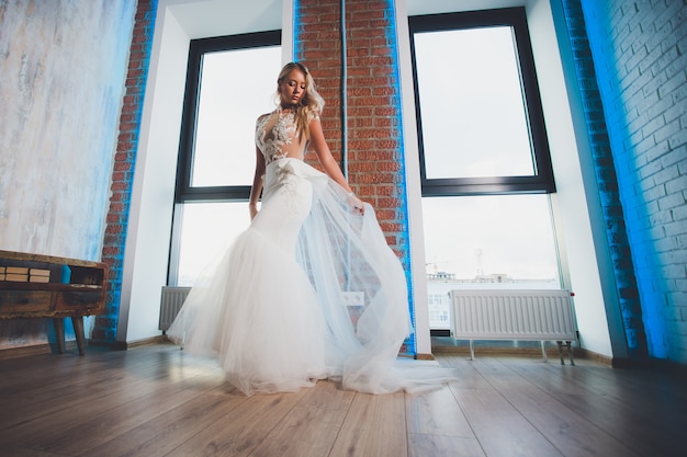 Belle femme blonde aux cheveux longs portant une robe avec des épaules nues, posant à la fenêtre.