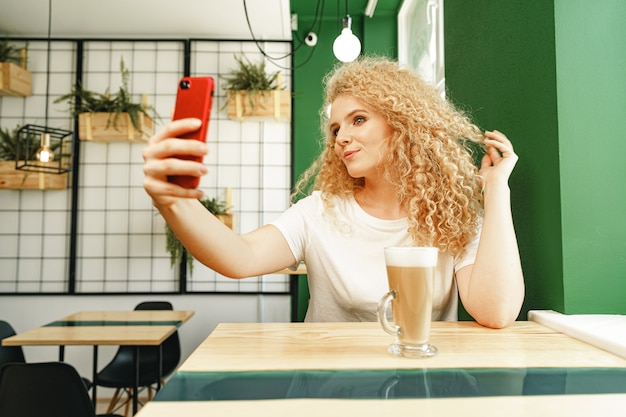 Belle femme blonde bouclée prenant selfie dans un café