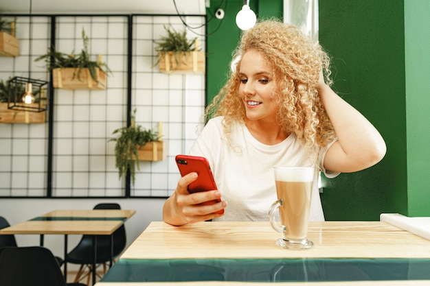 Belle femme blonde bouclée prenant selfie tout en étant assis à la table dans un café