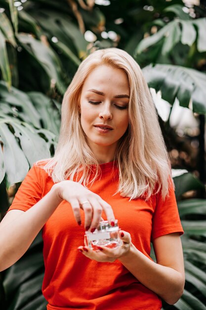 Belle femme blonde caucasienne ravie avec une peau propre et lisse, tenant un pot de crème, debout sur fond de forêt tropicale. Concept de cosmétiques naturels, produits de beauté et soins de la peau.