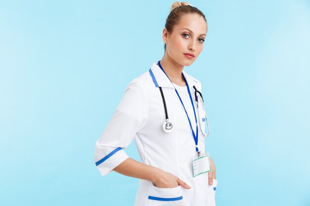 Belle femme blonde confiante médecin en uniforme debout isolé sur mur bleu