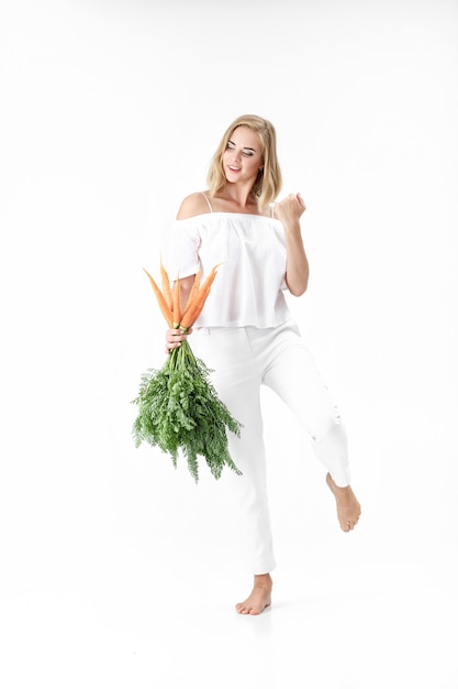 Belle femme blonde dans un chemisier blanc tenant une carotte fraîche avec des feuilles vertes sur fond blanc. Santé et alimentation