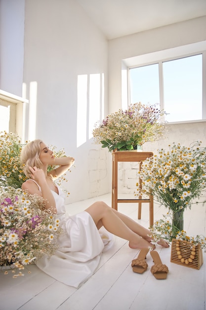 Belle femme blonde dans une robe blanche est assise sur le sol parmi les fleurs de camomille