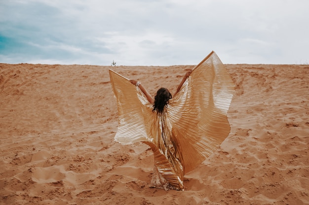 Une belle femme blonde dans une robe dorée avec des ailes brillantes, le costume danse une danse orientale orientale dans le désert sur le sable. Belle danseuse exotique dans un costume pour la danse du ventre.