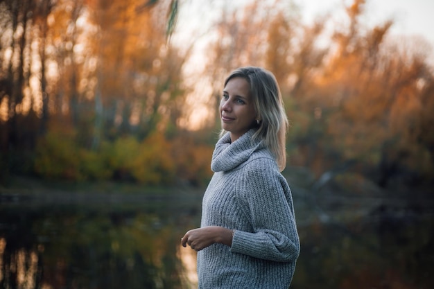 Belle femme blonde debout près de la rivière dans la forêt en détournant les yeux souriant et posant avec des arbres couverts de coucher de soleil en arrière-plan portant un chandail gris Promenade d'automne dans la forêt