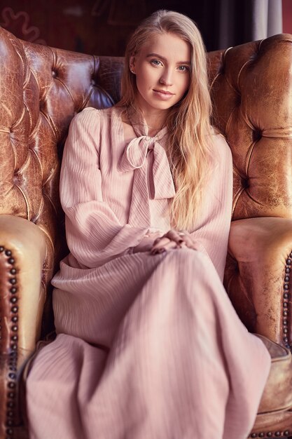 Belle femme blonde élégante en robe rose assis dans un fauteuil en cuir