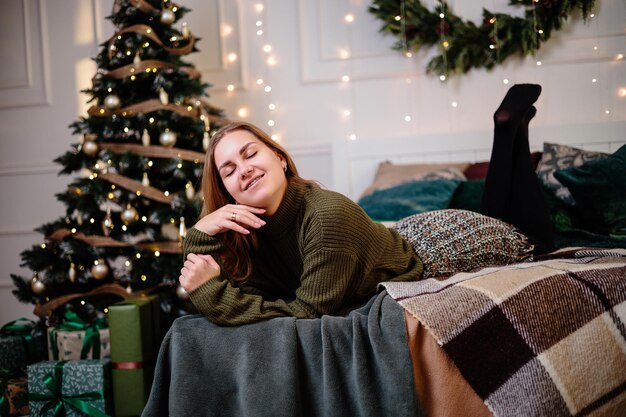 Une belle femme blonde est assise près d'un arbre de Noël dans une atmosphère du Nouvel An et se réjouit La femme est dans une chambre lumineuse et confortable joliment décorée pour Noël