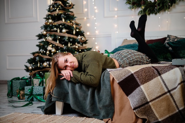 Une belle femme blonde est assise près d'un arbre de Noël dans une atmosphère du Nouvel An et se réjouit La femme est dans une chambre lumineuse et confortable joliment décorée pour Noël