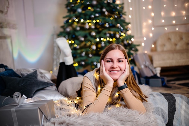 Une belle femme blonde est assise près d'un arbre de Noël dans une atmosphère du Nouvel An et se réjouit La femme est dans une chambre lumineuse et confortable joliment décorée pour Noël