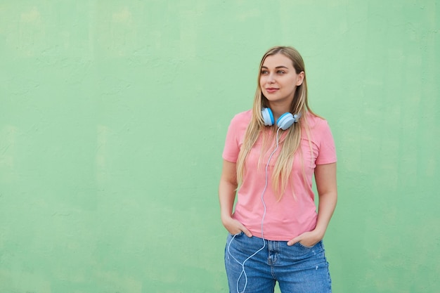 Belle femme blonde joyeuse avec un casque bleu dans un t-shirt rose se tient à l'extérieur contre un mur vert