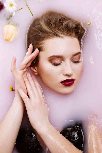 Belle femme blonde avec un maquillage parfait relaxant dans un bain avec du lait et des fleurs