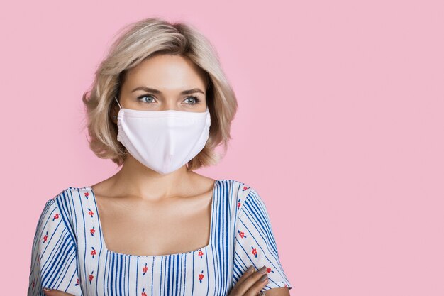 Belle femme blonde avec masque médical sur le visage posant sur un mur de studio rose avec espace libre vêtu d'une belle robe