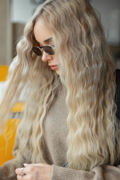 Belle femme blonde à la mode aux cheveux bouclés avec des lunettes de soleil élégantes dans un pull beige s'assoit et se peigne les cheveux