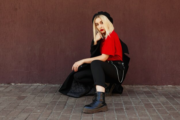 Belle femme blonde à la mode avec un béret noir avec un manteau d'automne noir posant dans la rue près du mur