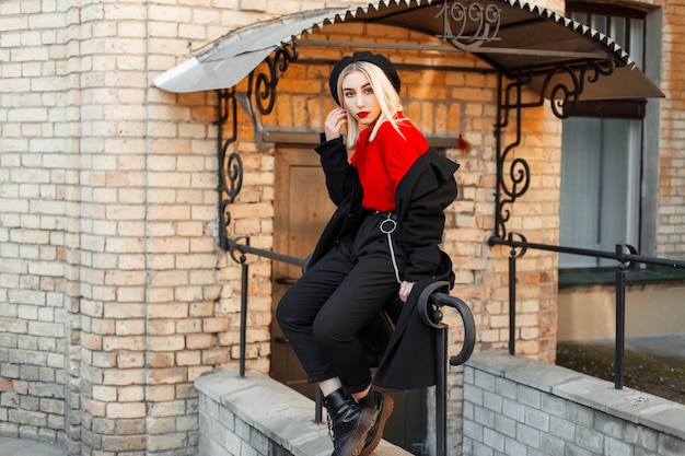 Belle femme blonde à la mode avec des lèvres rouges en manteau noir à la mode sur le porche