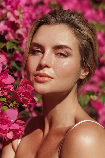 Belle femme blonde à la peau lisse et au maquillage naturel alors qu'elle pose sur fond de buisson aux délicates fleurs roses