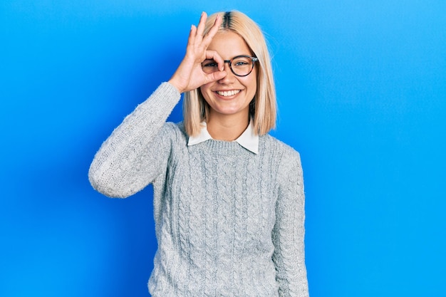 Belle femme blonde portant des lunettes faisant un geste correct avec la main souriante, les yeux regardant à travers les doigts avec un visage heureux.