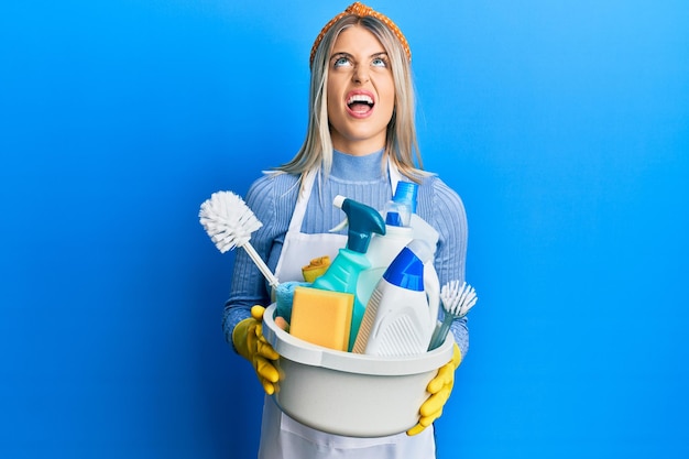 Une belle femme blonde portant un tablier nettoyant tenant des produits de nettoyage en colère et folle criant frustrée et furieuse criant de colère regardant en haut