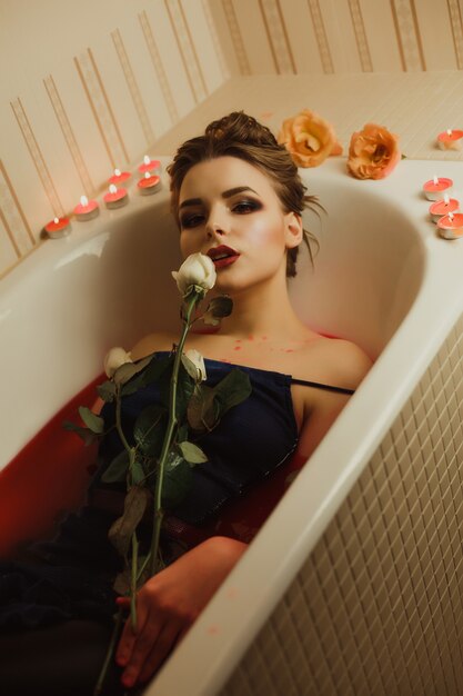 Belle femme blonde prend un bain avec de l'eau rouge et des bougies, tenant une rose