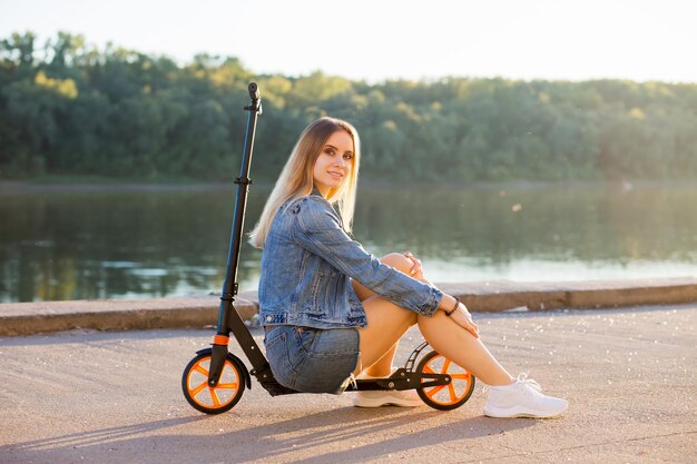 Belle femme blonde sexy en vêtements en denim est assise sur son scooter et sourit à la caméra