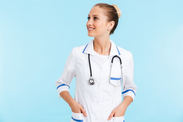 Belle femme blonde souriante médecin en uniforme debout isolé sur un mur bleu, en détournant les yeux