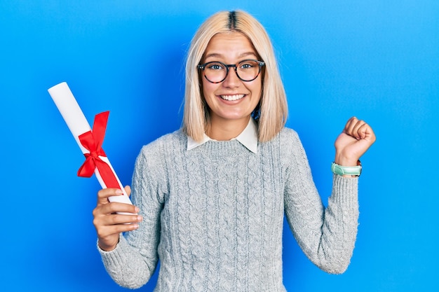 Belle femme blonde tenant un diplôme d'études supérieures criant fièrement, célébrant la victoire et le succès très excité avec le bras levé