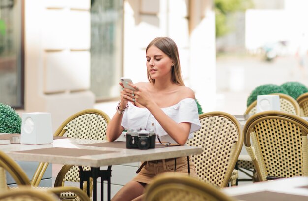 Belle femme blonde tenant le smartphone dans ses mains alors qu'il était assis à table dans le café de la rue