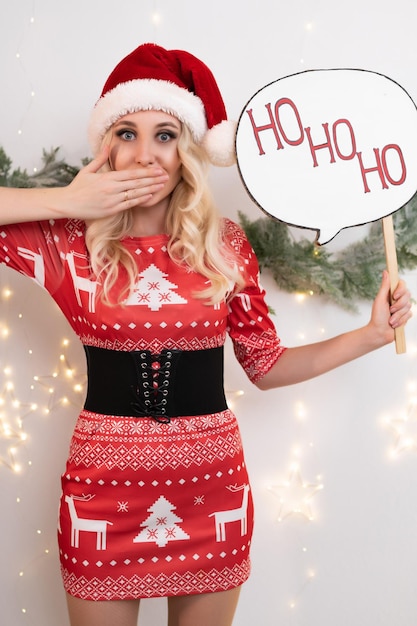 Belle femme blonde vêtue d'une robe de Noël rouge pose sur fond de branches d'arbres de Noël et d'une guirlande lumineuse Tient une pancarte dans sa main qui dit HoHoHo
