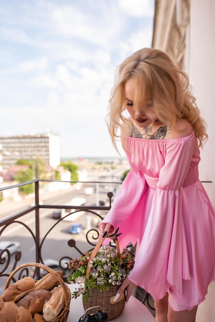 Belle femme blonde vêtue d'une robe sexy rose avec un tatouage sur la poitrine se dresse sur un balcon sur le fond de la ville soulève un panier de fleurs