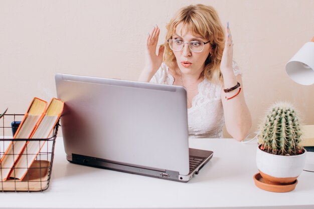 Une belle femme en blouse blanche est assise dans le bureau et regarde avec surprise le moniteur d'ordinateur portable