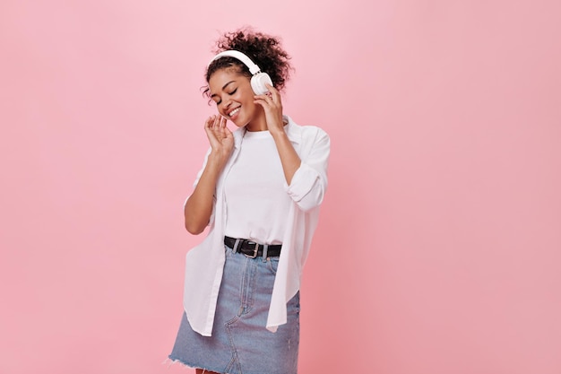 Belle femme de bonne humeur écoutant de la musique avec des écouteurs sur fond rose Portrait d'une fille bouclée heureuse en jupe en jean et chemise blanche appréciant la chanson sur fond isolé