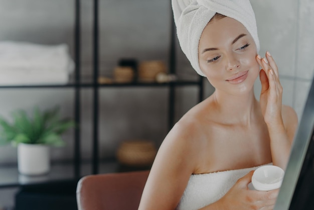 Belle femme en bonne santé applique une crème hydratante enveloppée dans une serviette de bain regarde attentivement la réflexion dans le miroir bénéficie d'un traitement de soins de la peau après la douche pose dans la salle de bain Concept de beauté