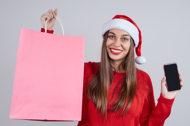 Belle femme en bonnet de Noel, pull rouge, retenez des paquets avec des achats et un téléphone mobile. Concept pour les ventes en ligne de Noël, commande, livraison.