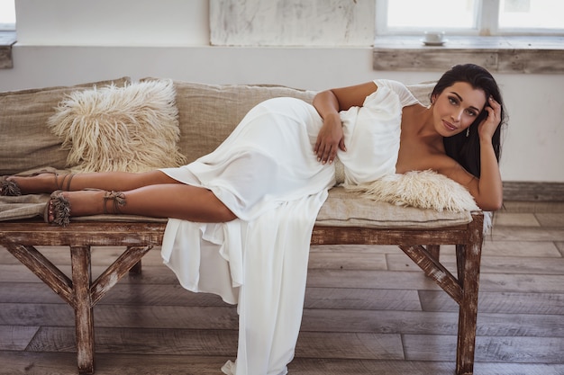 Belle femme bronzée, allongée sur le lit dans une robe blanche.