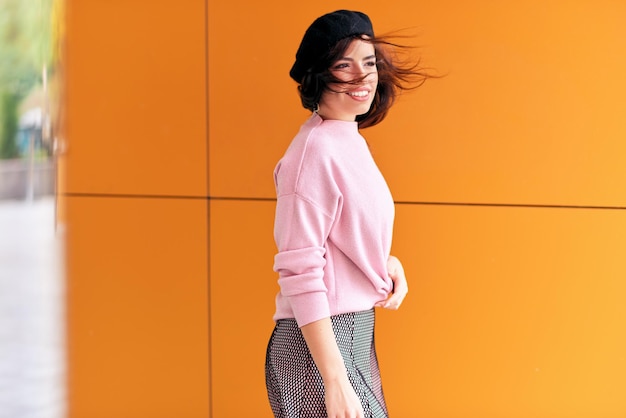 Belle femme brune aux cheveux venteux profitant du temps chaud de la ville Femme élégante moderne portant un pull rose et un béret chapeau noir contre un mur orange dans la rue de la ville Éclairage naturel