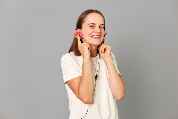 Belle femme brune en boutons de fleurs Tshirt blanc comme écouteurs écoutant de la musique avec plaisir en regardant la caméra avec une expression satisfaite isolée sur fond gris