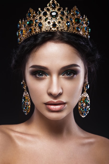 Belle femme brune avec une couronne d'or, des boucles d'oreilles et du maquillage de soirée professionnel