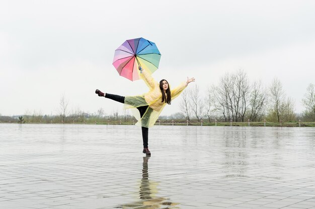 Belle femme brune en imperméable jaune tenant un parapluie arc-en-ciel sous la pluie