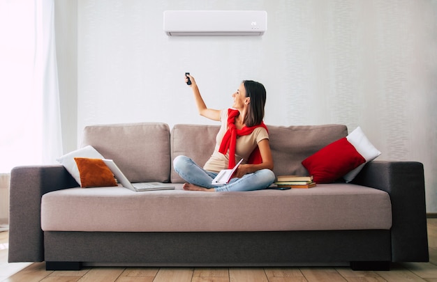 Belle femme brune moderne utilise le climatiseur assis sur le canapé et se reposant dans la chaude journée