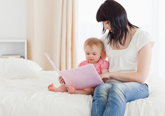 Belle femme brune montrant un livre à son bébé assis sur un lit