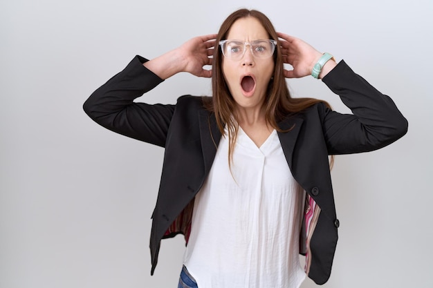 Belle femme brune portant une veste d'affaires et des lunettes folles et effrayées avec les mains sur la tête, effrayées et surprises du choc avec la bouche ouverte