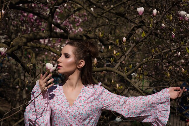 Belle femme brune posant près du magnolia en fleurs en robe à la mode