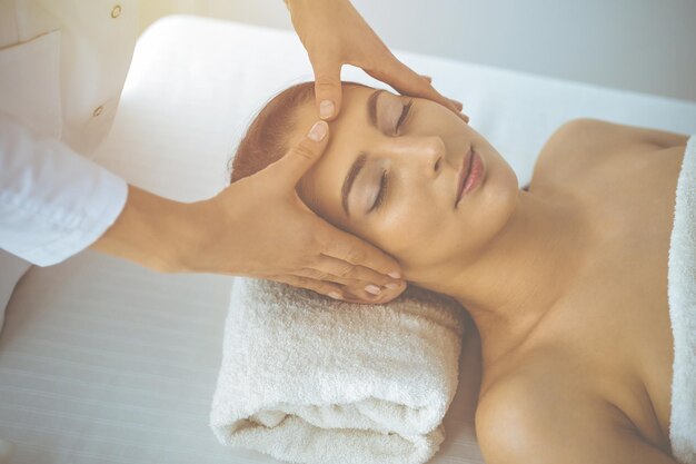 Belle femme brune profitant d'un massage du visage les yeux fermés dans un centre de spa ensoleillé. Concepts de soins relaxants et de médecine esthétique.