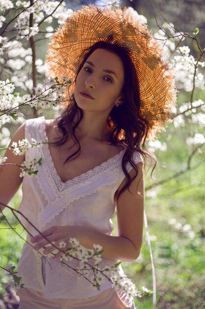 Belle femme brune se tient dans un verger de pommiers en fleurs dans un chapeau de paille et des vêtements roses