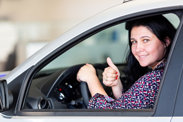 Belle femme brune souriante caucasienne assise dans la nouvelle voiture et montrant les pouces vers le haut.